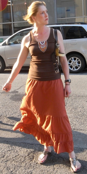Jessica stands, right arm outstretched, looking left into the distance. She wears an orange skirt with sandals and a brown tank top with a small pattern in the center. She is lit from the left which highlights her auburn hair held up by an ornamental hair tie.